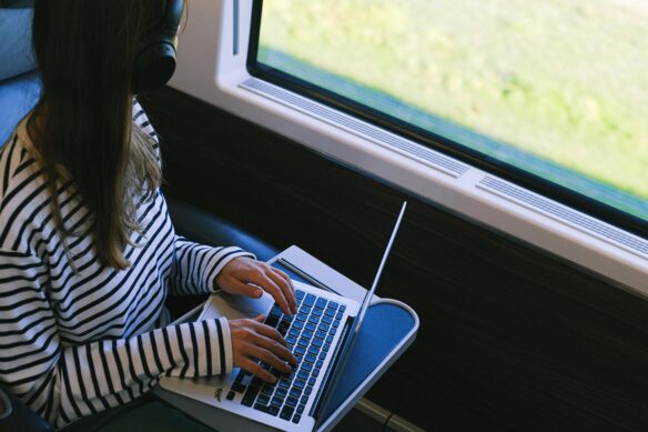 A woman looking out the window of a train while typing on her laptop.