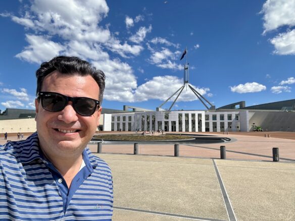 Alex in front of the Parliament House in Canberra with a partly cloudy blue sky overhead. 