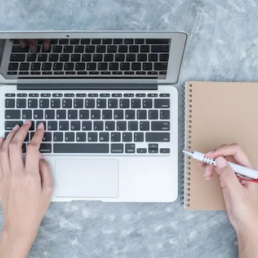 Someone with an open laptop with one hand on the keyboard and another holding a pen over a notebook.