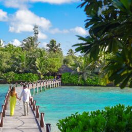 A couple holding hands while walking on a wooden bridge over turquoise water.