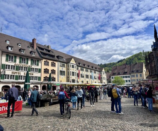 Many shoppers perusing the stalls at Muenstermarkt in Muensterplatz in Freiburg, Germany on a beautiful day with a partly cloudy blue sky.