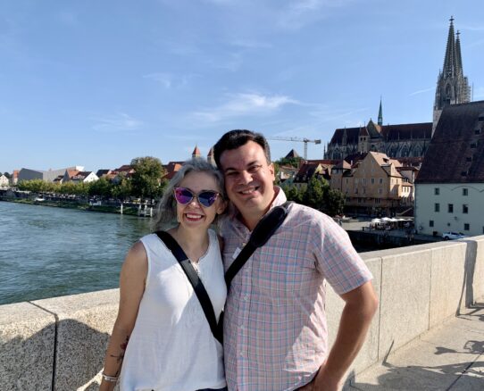 Alex and Bell of Wanderlust Marriage atop the Stone Bridge in Regensburg, Germany with a blue sky above.