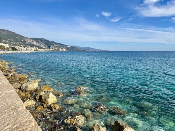The rocky coastline of the French Riviera with crystal clear turquoise water with Menton and the mountains in the background.