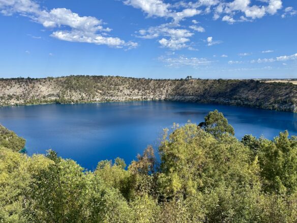 Tall trees surround Blue Lake in Mount Gambier, South Australia with a partly cloudy blue sky above